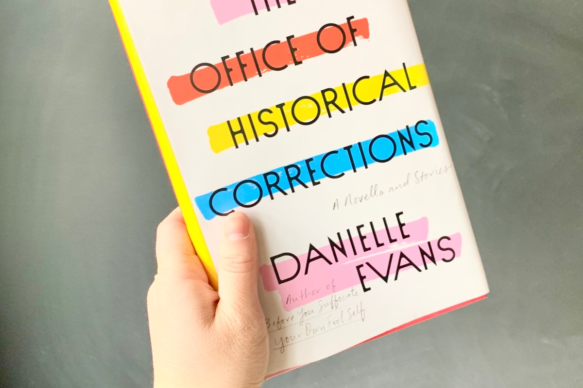 A copy of The Office of Historical Corrections by Danielle Evans being held up against a chalkboard wall.